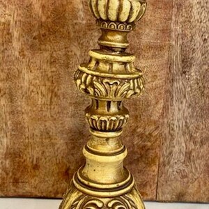 May include: A gold-toned candlestick holder featuring a wide, scalloped top rim, a multi-tiered central column with intricate ribbed and floral detailing, and a broad, triangular base adorned with ornate, symmetrical scrollwork and relief patterns.
