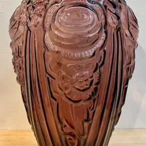 Puede incluir: Un jarrón de cristal color amatista vintage con un diseño Art Déco. El jarrón presenta patrones florales y geométricos en relieve, con una parte superior ancha y una base cónica. El cristal tiene un acabado brillante y el jarrón probablemente se utiliza con fines decorativos.