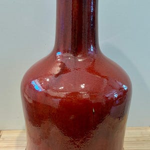Pode incluir: Um vaso de cerâmica vermelho brilhante, de forma alongada, com uma base larga e um pescoço estreito. A superfície é texturizada e a borda superior é escura. A base apresenta uma borda decorativa ondulada. O vaso é provavelmente usado para fins decorativos.