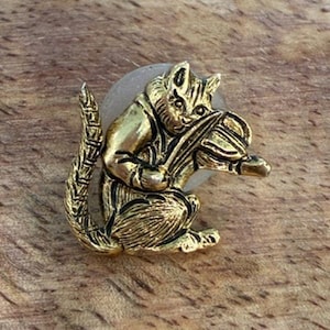 May include: A gold-toned metal lapel pin featuring a cat playing a violin. The cat is depicted in a seated position with its tail curved upward, holding a violin and bow. The design includes detailed etched lines to show the cat's fur texture.