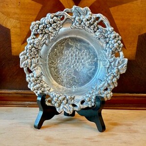 May include: A round silver-toned metal bowl featuring an intricate embossed grape and vine motif along the scalloped rim and a central grape cluster design. The bowl is displayed on a black three-legged stand for upright presentation.
