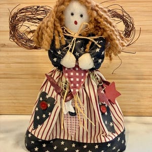 May include: A patriotic doll with a fabric body, featuring a red, white, and blue color scheme. The doll has a star-patterned skirt, a navy blue top with white stars, and a red and white striped skirt. It has brown yarn hair and a twig halo.