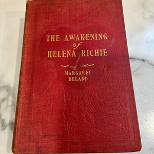 Könnte beinhalten: Ein altes Buch mit dem Titel "The Awakening of Helena Richie" von Margaret Deland. Das Buch hat einen roten Einband mit goldener Schrift und geprägten floralen Mustern. Der Einband des Buches weist Gebrauchsspuren auf.