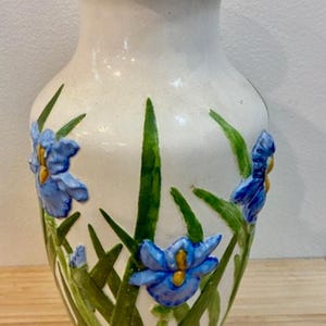 May include: A white ceramic vase with a flared top and base, adorned with hand-painted blue iris flowers and green leaves. The vase is approximately 8 inches tall, a decorative home accent.