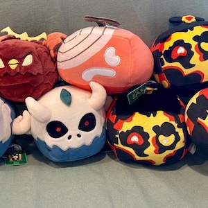 Könnte beinhalten: Eine Sammlung von Plüschtieren in verschiedenen Formen und Farben. Die Plüschtiere umfassen einen rosa Apfel, eine rote Eule, eine orangefarbene Zwiebel und mehrere schwarz-gelb gemusterte Figuren. Einige haben Hörner oder Stiele.