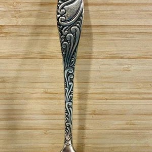 May include: Silver ladle with ornate, swirling floral design on the handle. The ladle has a rounded bowl and is set against a light brown wooden background. A classic kitchen utensil.