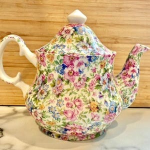 May include: A floral patterned teapot with a white handle and spout. The teapot features a lid with a white finial. The floral pattern includes pink, blue, and yellow flowers on a cream background. The teapot is made of ceramic.
