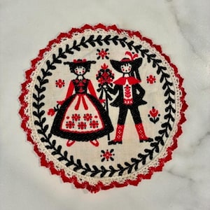 May include: A round fabric doily featuring black and red embroidery of two figures in traditional attire. The design is enclosed by a black leaf border and a scalloped red and white crocheted edge on a cream-colored background.