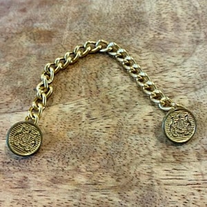 May include: A gold-toned metal cardigan clip featuring a short curb chain connecting two round buttons. Each button displays a detailed embossed crest design on its surface, providing a secure and decorative closure for garments.