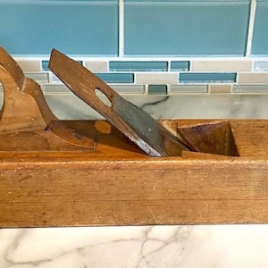 May include: A vintage wooden hand plane, featuring a curved handle and a metal blade. The plane is constructed from light-coloured wood, displaying signs of use. The blade is partially visible. This woodworking tool measures approximately 50 cm in length.