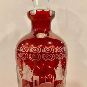 May include: A ruby red glass bottle with a clear, faceted stopper. The bottle features etched scroll and landscape designs. A small, oval sticker is visible on the bottle. The bottle is likely used for perfume or decorative purposes.