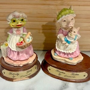 May include: Two ceramic figurines: a frog holding a basket and a pig holding a piglet. Both are dressed in pink and white aprons and bonnets. The figurines sit on wooden bases with nameplates that read "Felicity by Leonardo" and "Heather Footer by Leonardo."