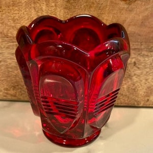 May include: A ruby red glass vase with a scalloped rim and geometric design. The vase has a rounded base and a series of vertical, rectangular panels with horizontal lines. The glass is transparent, allowing light to pass through.
