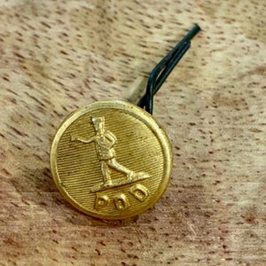 Peut inclure: Un bouton rond en métal doré présentant un relief d'une personne en uniforme marchant vers l'avant. Sous la silhouette, les lettres P O D sont gravées sur une ligne horizontale. Une boucle en fil de fer vert foncé est fixée au dos.