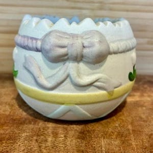 May include: A ceramic egg-shaped container with a light blue and white color scheme. The container features a bow, a yellow band, and green floral accents. The top edge has a scalloped design.