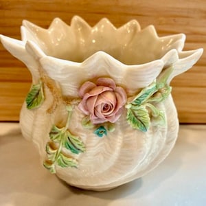 Könnte beinhalten: Cremefarbene Keramikvase mit gewelltem Rand und geripptem Körper. Eine rosa Rose und grüne Blätter sind auf der Vorderseite angebracht. Die Vase hat ein dekoratives, florales Design.