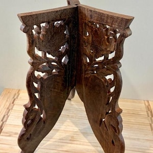 May include: A dark brown wooden stand featuring two interlocking panels with intricate cut-out floral and leaf carvings. The structure is composed of two vertical pieces joined at a central axis, creating a stable base for displaying objects.