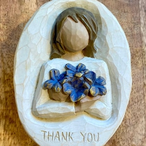 Könnte beinhalten: Eine geschnitzte, cremefarbene Reliefskulptur einer Figur, die blaue Blumen hält. Die Figur hat braunes Haar und trägt ein weißes Kleid. Die Worte "THANK YOU" sind am unteren Rand der ovalen Skulptur eingraviert.