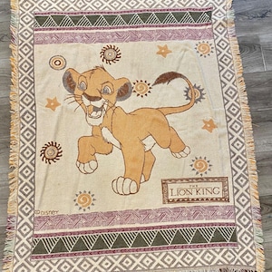 May include: A tan throw blanket featuring a cartoon image of Simba from The Lion King. The blanket has a decorative border with geometric patterns and the words "The Lion King" in a brown rectangle.