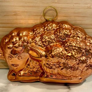 May include: A copper-colored sheep-shaped mold with a textured surface and a small loop for hanging. The mold is designed with detailed features, including the sheep's face, ears, and woolly body. The mold is likely used for baking or decorative purposes.