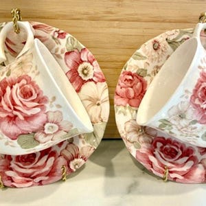 Puede incluir: Dos tazas de té y platillos blancos con un estampado floral de rosas en tonos rosa y crema. Las tazas de té están suspendidas sobre los platillos mediante ganchos dorados. Los platillos y las tazas están decorados con un diseño floral repetido.