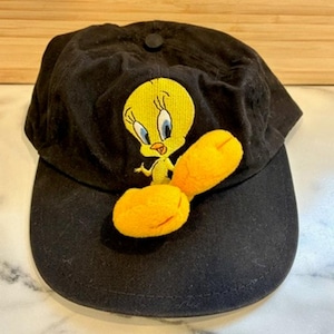 Puede incluir: Gorra de béisbol negra con un bordado del personaje amarillo Piolín, con ojos azules y pies naranjas. La gorra tiene una visera curva y un botón en la parte superior. Un accesorio divertido.