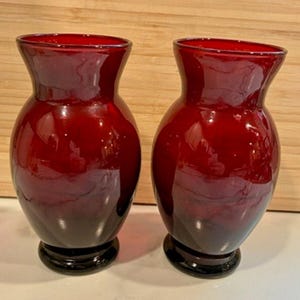 May include: Two ruby red glass vases with a glossy finish. The vases have a rounded body, a flared neck, and a black base. The vases are displayed on a white surface with a wooden background.