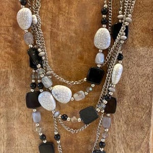 May include: A multi-strand necklace featuring silver-tone metal chains interspersed with black square beads, white crackle-patterned oval beads, and smaller translucent grey beads. The layered design creates a cascading effect with varied bead shapes and textures throughout the piece.