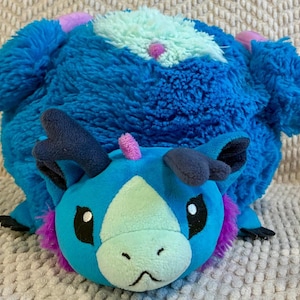 May include: A plush toy in the shape of a blue creature with a round body and a dragon-like head. The head is teal with purple and pink accents, black eyes, and dark blue horns. The body is a darker shade of blue with a fluffy texture.