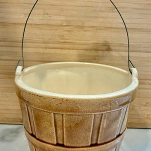 May include: A brown ceramic bucket with a wire handle and a wooden grip. The bucket has a basket weave design and a cream-colored interior. The handle is attached to the bucket with metal wire.