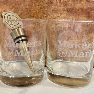 May include: Two clear Maker's Mark whiskey glasses with a silver-colored bottle stopper. The glasses have a rounded base and the Maker's Mark logo etched on the front. The bottle stopper is inserted into one of the glasses.