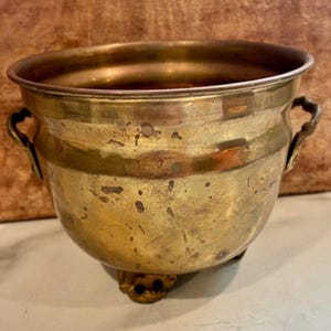 May include: A round brass planter featuring a weathered, golden-toned surface with visible patina and dark spots. The vessel includes two small, decorative side handles and rests on three ornate, sculpted feet, providing a stable base.