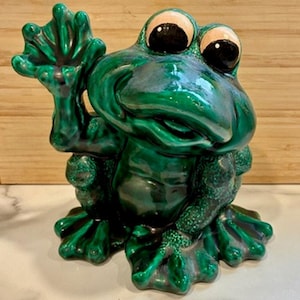 Może przedstawiać: Ceramiczna figurka żaby w błyszczącym szmaragdowym kolorze. Żaba jest przedstawiona w pozycji siedzącej, z jedną ręką uniesioną w geście machania. Żaba ma duże, wyraziste oczy i szczegółowe stopy. Przedmiot dekoracyjny.