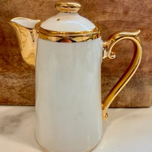 Op de afbeelding: Een witte keramische koffiepot met gouden details en een handvat. De pot heeft een deksel met een gouden knop en een tuit. De handgreep is gebogen.