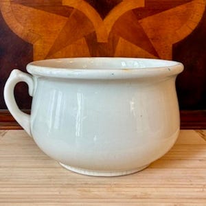 Può includere: Un vaso da notte in ceramica bianca caratterizzato da un corpo liscio e arrotondato con un'unica maniglia curva sul lato sinistro. Il recipiente presenta un bordo largo e svasato e un interno profondo, con una finitura lucida esterna.