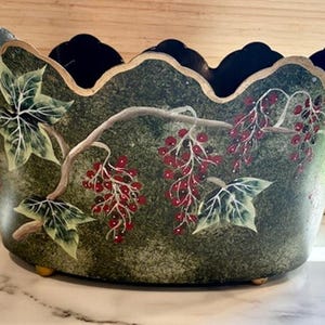 May include: A decorative bowl with a scalloped rim and a dark green mottled finish. The exterior features a hand-painted design of green ivy leaves and clusters of small red berries on light brown branches. The bowl rests on small gold-toned feet.