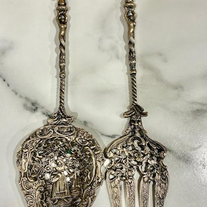 May include: Ornate silver serving set featuring a decorative spoon and fork. The spoon has a detailed floral design with a central figure, while the fork has three tines. Both pieces have twisted handles and fleur-de-lis tops.