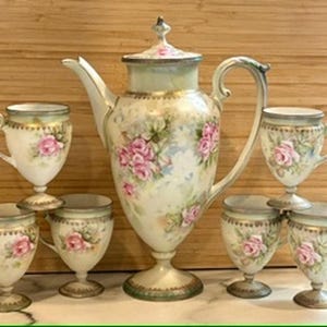 May include: A vintage porcelain tea set featuring a large teapot and six matching cups. The set is decorated with pink roses and green leaves on a cream-colored background. The teapot has a lid and a curved handle.