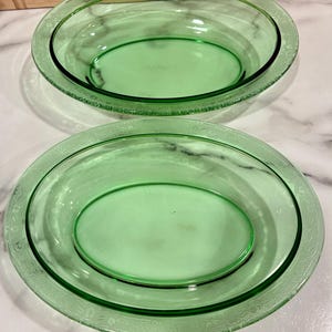 Könnte beinhalten: Zwei grüne Glasservierschalen. Die Schalen haben eine ovale Form mit einem dekorativen, geätzten Rand. Die Schalen sind auf einer weißen und grauen Marmoroberfläche verschachtelt. Die Schalen sind leer.