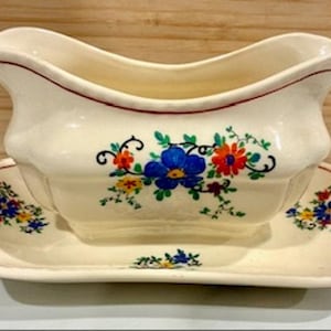 Könnte beinhalten: Cremefarbene Sauciere und passende Untertasse. Das Boot hat einen gebogenen Griff und eine rote Zierleiste. Blumendesigns in Blau, Orange und Gelb verzieren das Boot und den Teller. Der Hintergrund ist eine hellbraune Holzoberfläche.