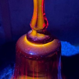 May include: A glass bell with a rounded base and a long, slender handle. The bell is primarily yellow with red accents, set against a dark blue background.