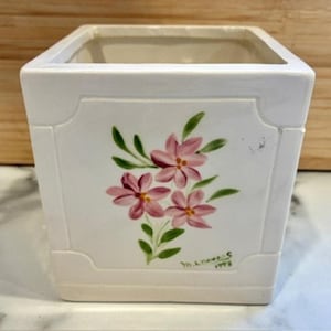 May include: A square, white ceramic planter with a floral design. The planter features a hand-painted cluster of pink flowers with green leaves. The artist's signature and the year 1998 are visible on the front. The planter has a rectangular shape.
