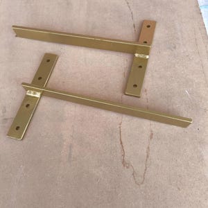 May include: Two gold-colored metal shelf brackets. Each bracket has a horizontal bar and a vertical support with pre-drilled holes for mounting. The brackets are designed to support shelves and add a touch of elegance to any space.
