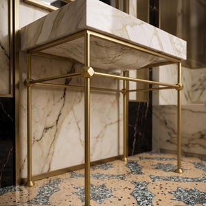 Puede incluir: Un lavabo rectangular de mármol con patas de metal dorado. El lavabo tiene un patrón de mármol blanco y beige. Las patas están conectadas por barras horizontales. El baño tiene paredes de mármol y un suelo estampado.