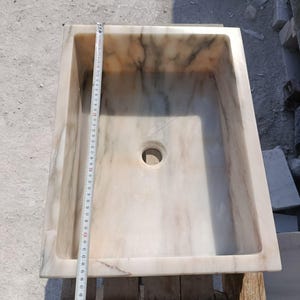 May include: Rectangular marble sink with a central drain hole. The sink has a light beige and brown veined pattern. A measuring tape shows the sink is approximately 21 inches long.