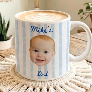 May include: A white ceramic mug with blue vertical stripes, featuring a baby's face and the text "Mike's Dad." The mug sits on a cream-colored macrame coaster, filled with a latte.