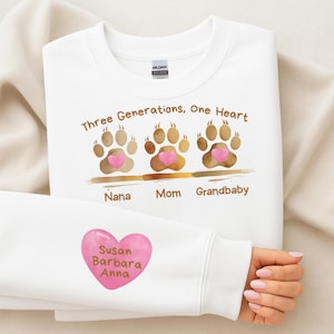 Peut inclure: Sweat-shirt blanc avec l'inscription "Three Generations, One Heart" et des empreintes de pattes avec des cœurs roses. La manche présente un cœur rose avec les noms "Susan Barbara Anna".