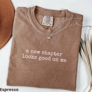 Op de afbeelding: Espresso-kleurig Comfort Colors T-shirt met de witte tekst "a new chapter looks good on me". Een zilveren ketting met een hanger ligt over het shirt. Een bruine hoed is linksboven zichtbaar.