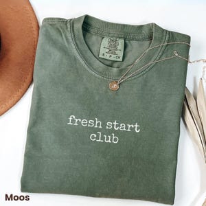 May include: An olive green t-shirt with the text "fresh start club" in white. A gold necklace with a small pendant rests on the shirt. A brown hat is partially visible in the upper left corner.