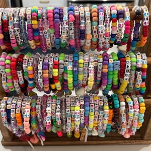 May include: A wooden display case filled with colorful beaded bracelets. Each bracelet features a variety of colors and lettered beads spelling out words and phrases. The bracelets are arranged in rows, showcasing a diverse collection of handmade accessories.